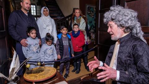 A man in a grey curly wig and velvet coat speaks to some children while looking at astronomical equipment