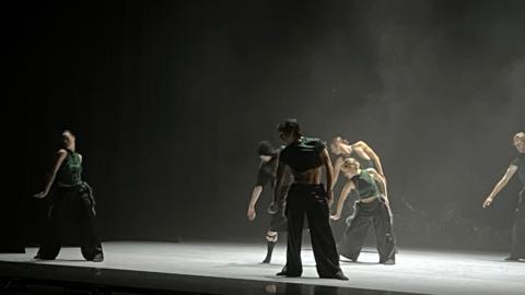 Six dancers on a stage. They are wearing green tops and black bottoms and are learning towards stage right. 