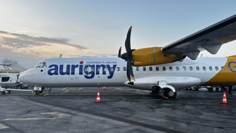 The picture shows an Aurigny aircraft parked on an airport apron. The plane has two large propellers, one of which is clearly visible near the wing. The fuselage is white with the word “aurigny” written in bold blue letters, and underneath it reads “Guernsey’s Airline.” The tail section and engine housing are painted in a bright yellow colour. There are orange traffic cones placed near the aircraft, and the ground appears to be a tarmac surface. In the background, there is airport equipment and a partly cloudy sky.