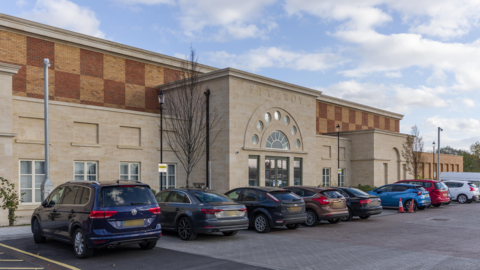 A large, modern, cream-coloured brick building. There are cars parked in front of it. It has a portico reception area. The upper storey has orange and brown-coloured bricks.