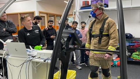 A firefighter is standing on a large treadmill-type machine, with a full beige and black firefighter suit on, as well as an oxygen mask that is strapped around his head and goes over his mouth and nose. He is wearing black glasses and is looking over a group of people who are in black, watching him next to a laptop.