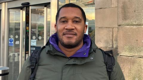 Simon Coombs stood outside Sheffield train station. He has short, dark hair and is wearing a green jacket with a purple sweater underneath.