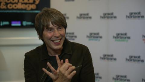 Professor Brian Cox sits in front of a white background. He wears a black jacket and a black T-shirt. He has collar-length dark hair and a sweeping fringe.