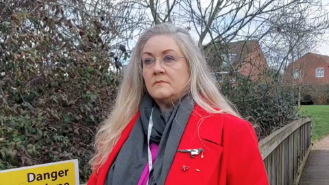 Julie Stevenson who has long grey/blonde hair and is wearing rimless glasses, a red coat over a grey scarf and pink top. She is standing on a wooden bridge with houses glimpsed behind her through trees/shrubs.