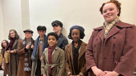 Eight children in 1940s costumes standing in a hallway.