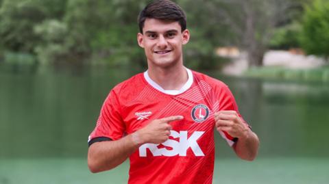 Rob Apter wearing a red Charlton Athletic kit, pointing at the club crest with his right hand and holding his shirt with his left hand, standing in front of a lake