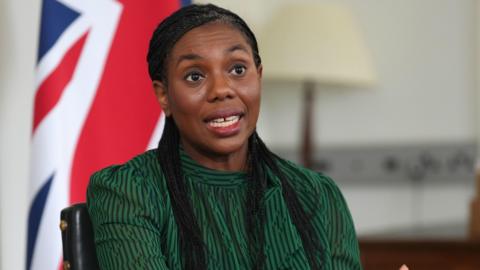 Kemi Badenoch in a green dress and with long platted black hair sitting in front of the union flag.