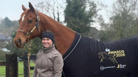 A brown horse wearing a horse coat that reads 'RoR Awards 2026'. The woman is standing next to the horse wearing a navy woolly hat and a grey fleeced jacket. She is smiling. Behind them is a wooden fence and a field.