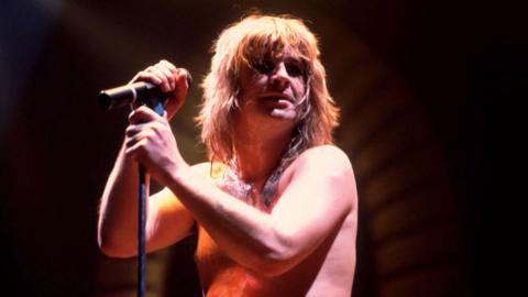 Ozzy on stage in 1982, with no top on, in front of a mic.