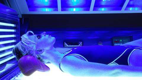 A lady lies inside a sunbed with a strong blue light. She has goggles and a swimming costume on and we can see her head to the top of her legs