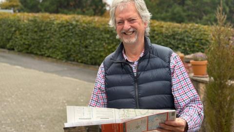 A man in a blue padded gilet with a checked shirt underneath and a grey beard and long grey hair. He is holding a map and smiling in front of a hedge and driveway.
