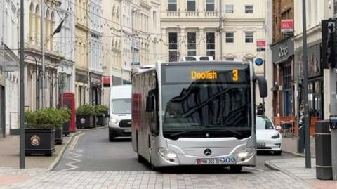 A bus with the number three, which says Doolish on it, is driving down a busy Manx street.