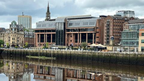 Newcastle Crown Court reflected in the River Tyne running in front of it. It is an imposing building made from smooth red stone with massive black windows and tall columns along its frontage.