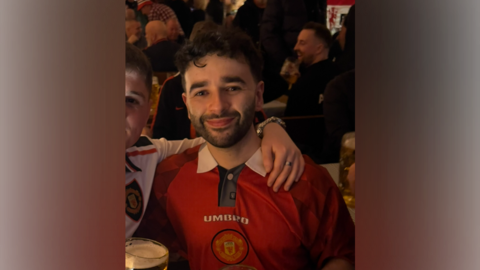 A man with black, curly hair, and a beard, looks directly at the camera. He is wearing a red football shirt bearing the word 'Umbro', and a football emblem underneath. Another man has his arm around his shoulder. Crowds can be seen behind them. 