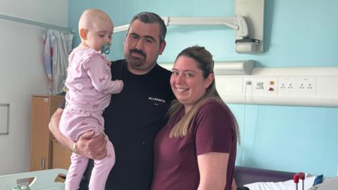 Avarni-Mae being held by her dad Daniel, who is standing next to Chanelle. Avarni-Mae is looking down and is wearing pink pajamas with bows and has a blue dummy in her mouth. Daniel has grey, short hair and a dark beard. He is wearing a black t-shirt. Chanelle has brown long hair and is wearing a purple top. Both are looking into the camera and smiling. They are standing in a hospital room with a light blue wall.