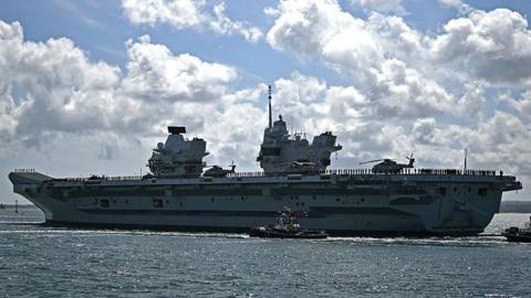 British aircraft carrier HMS Prince of Wales as it leaves Portsmouth Naval Base. It is during the day, the ship takes the entire picture. The waters are calm and the sky is blue but full of clouds.