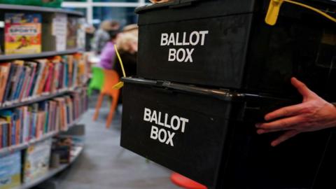 A person carries two ballot boxes stacked on top of one another through a library.