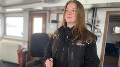 Ellie stands behind the wheel of the ferry, looking out to sea. She has brown hair and blue eyes and is wearing a black zip-up fleece.