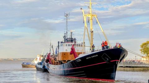 A fishing trawler, painted black and white with a red stripe along the length of the hull, is pulled by a tug boat along the flat calm Humber Estuary.