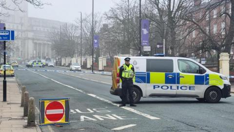 A road in Leeds which has been closed to traffic. A police van is parked across the road with an officer in uniform stood near it
