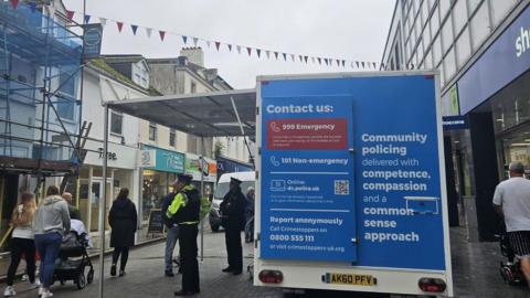 Devon and Cornwall Police's mobile police station parked in a town centre. The van is blue and white. A police officer is stood in front of the mobile station. A woman dressed in blue jeans and a grey hoodie is walking past the van.