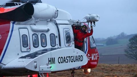 A man in a red bodysuit and helmet leaning out the side of a HM Coastguard helicopter in white and red.