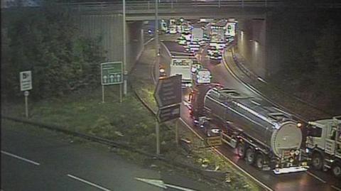 A roundabout with traffic queuing on it, including cars, lorries and a tanker.