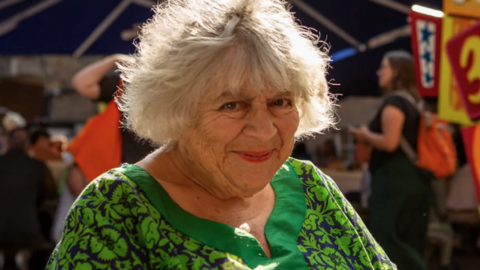 Miriam Margolyes, an older lady with grey bobbed hair, smiles at the camera in the sunshine in Edinburgh, wearing a green floral dress
