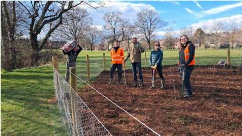 A park with grass and trees to the left. There is a fenced off area containing soil on which four people are standing -two have orange hi-vis vests. The three are holding spades. Another person is carrying a bag of compost on his shoulder and is on the other side of the fence.