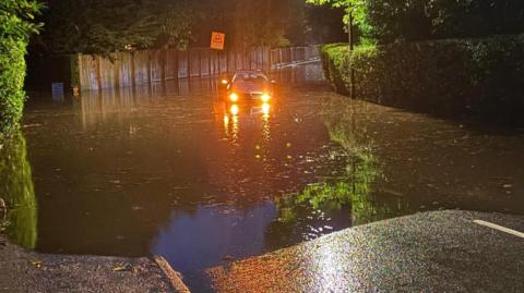 A car, with its headlights on, struggles through water in a flooded street