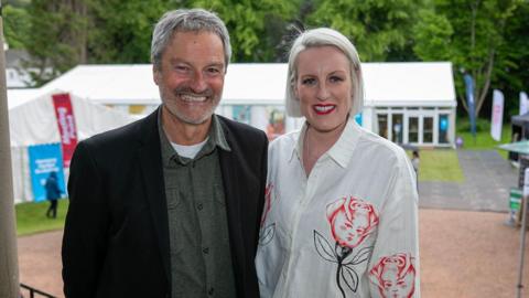  Gavin Esler and Steph McGovern - he has grey hair and grey stubble and is wearing a dark jacket with a green shirt underneath. She has blonde hair and is wearing a white blouse with a rose pattern on it.