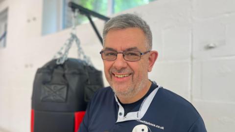 Peter Martin stands in a gym with kick boxing gear behind him. He is wearing glasses and a blue top, has short greying hair and stubble and is smiling at the camera.