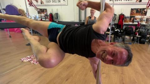 Paul Bradley wearing a black sports vest and shorts holding on to a vertical metal bar under his arms while able to hold his body horizontal. His neck and arm muscles look strained but he has a smile on his face.