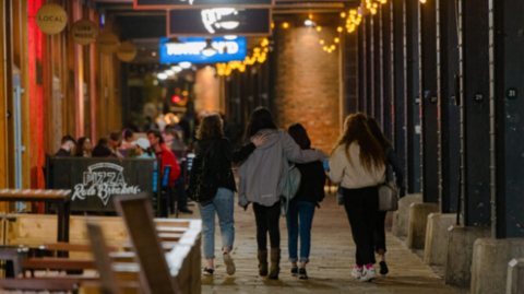 Four young women out at night together with bars and restaurants in the background alongside the Watershed on the harbourside in Bristol