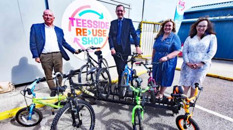(L-R) Councillor Peter Gavigan, SUEZ Regional Director Steve Patterson, Councillor Lisa Evans and Amanda Padfield, director of public sector development at SUEZ. All four are standing in front of a small bike rack which is supporting seven children's bicycles. A sign on the wall marks the site as the Teesside Reuse Shop.