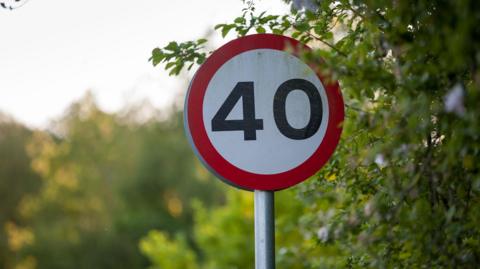 A 40mph road sign, it is a red circle around a white background with the number 40 in black numerals. There is a hedgerow to the right and the photo was taken on a sunny day.