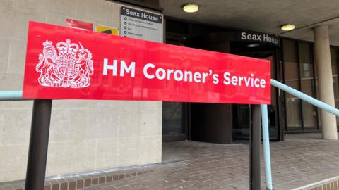 The close up of building with a red sign that reads HM Coroner's Service.
Behind is the doors of the building