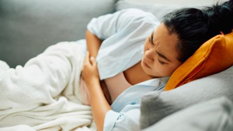 A woman lies on a sofa at home in visible discomfort with her arms on her stomach.