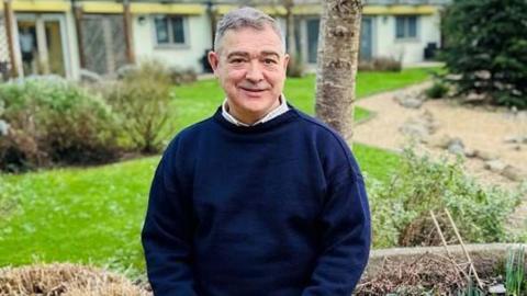 Les Bourgs Hospice chief executive Rob Jones sitting in a garden with a tree and a large grass lawn behind him. He is wearing a dark blue jumper and is smiling.