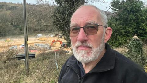 Brian Garlick. He has short grey hair and facial hair. he is pictured outside and there is a construction site in the distance behind him. He is wearing glasses and a black fleece with a car embroidered on the chest. He is looking at the camera and smiling slightly 