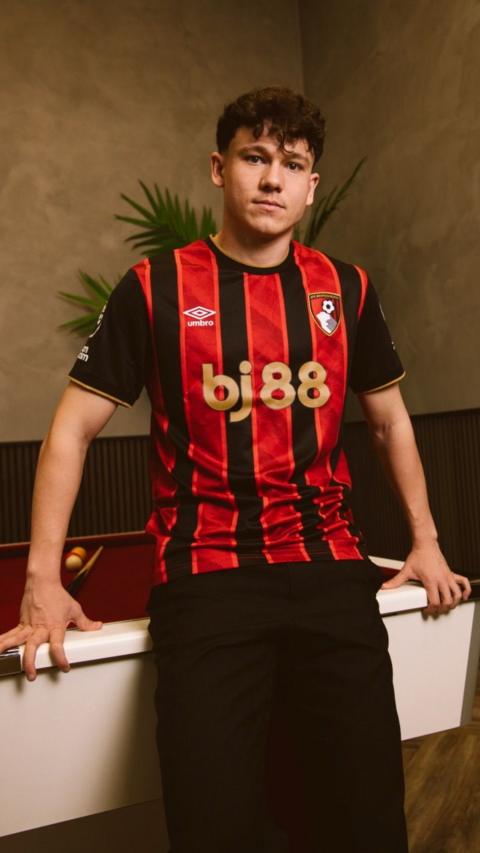 Alex Toth is leaning against a white desk wearing the Cherries red and black striped kit.