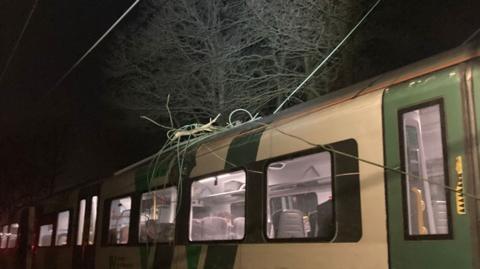 A green and cream train pictured at night, with wires tangled on its roof. The train lights are on and a passenger can just be seen sitting inside.