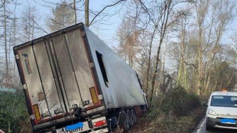 A large lorry, blackened by fire, tilted to one side on a muddy grass verse in a wooded area beside the road. There is a large hole in one side of the lorry. A highways van with an orange light on top is parked by the road next to the vehicle.
