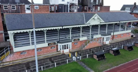 A Victorian era football stand with pastel green paint and tiled roof can be seen, with houses behind it and grass to the front.