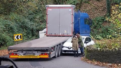 A large lorry with a flat back on the left stuck on the corner of a road with a small white car wedged between the lorry and a wall on the right. There are bushes on either side of the road. The front of the lorry is blue. 