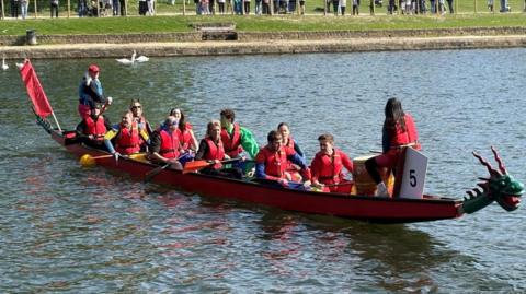 A long dragon boat moving across a calm body of water on a bright, clear day. The boat is painted primarily in red and carries a group of paddlers seated in pairs along its length, all wearing red life jackets and holding paddles that are resting or raised slightly above the water. At the front of the boat, a decorative dragon head is attached, and a small white number sign marked “5” is visible near the bow.