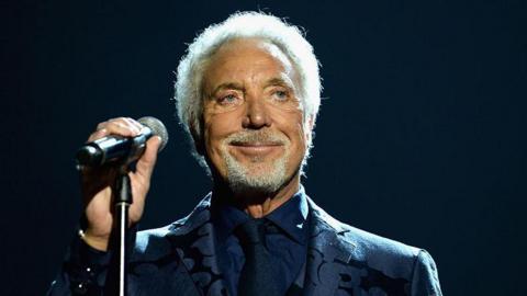 Sir Tom Jones holds a mic stand and grins