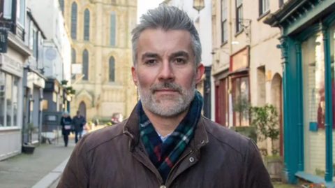 York and North Yorkshire Mayor David Skaith is looking straight at the camera. He is wearing a brown jacket and a scarf. York Minster is in the picture's background.