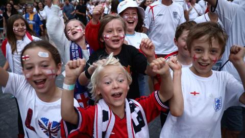 Young England fans, boys and girls, in red and white colours, cheering with their arms in the air.