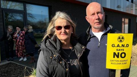 Protesters outside council offices in Horncastle. A man with short hair is holding a placard which reads: Gayton says no - Gayton against nuclear dump. A woman wearing a black coat and sunglasses stands next to him.